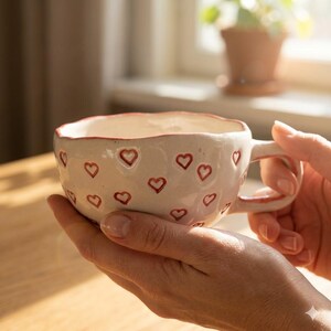 Könnte beinhalten: Eine weiße Keramiktasse mit rotem Rand und roten Herzmotiven. Die Tasse hat einen Henkel und wird mit zwei Händen gehalten. Die Tasse steht vor einem Fenster mit einer Pflanze im Hintergrund.