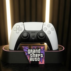 Può includere: Un controller per videogiochi bianco e nero appoggiato su una base di ricarica nera. La base presenta un emblema grafico viola e rosa con il testo Grand Theft Auto VI, che mostra sagome di palme dietro le lettere bianche in grassetto.