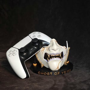 Op de afbeelding: Een wit en zwart videogame-controller naast een wit samurai-achtig gezichtsmasker met gouden accenten en ontblote tanden. Het masker rust op een ronde zwarte voet met de gouden tekst GHOST OF YOTEI gedrukt op de voorste rand.