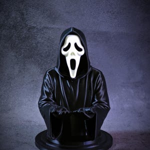 May include: A black bust of the Ghostface killer from the Scream horror film franchise. The figure wears a black hooded robe and a white mask with a screaming expression. The bust is on a black circular base.