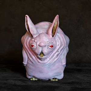 Pode incluir: Uma pequena estatueta arredondada de uma criatura com pele rosa pálido, orelhas pontiagudas proeminentes e olhos vermelhos brilhantes. A figura apresenta um corpo largo e robusto, um nariz pequeno e um dente amarelo visível na boca.