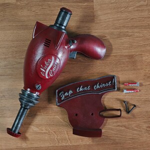 Puede incluir: Una réplica de utilería roja y negra de un arma Nuka Cola, con las palabras "Nuka Cola" en el lateral. La utilería va acompañada de un letrero a juego que dice "Zap that thirst!" y accesorios de montaje.