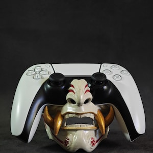 May include: A white and black video game controller with a custom faceplate featuring a detailed, off-white Oni mask with red and gold accents. The mask is centered on the controller, adding a unique aesthetic.