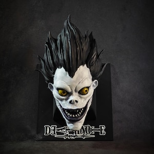 Può includere: Una scultura da parete del personaggio Ryuk di Death Note, caratterizzata da un volto pallido, capelli neri a punta e occhi gialli penetranti. La figura è montata su una placca rettangolare nera con il logo Death Note stampato in bianco.
