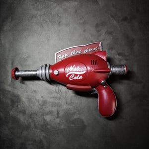 Könnte beinhalten: Eine rote Strahlenpistole mit dem Nuka-Cola-Logo an der Seite und einem rechteckigen Schild dahinter mit der Aufschrift Zap that thirst!. Das Gerät hat einen metallisch silbernen Lauf, einen geschwungenen Griff und eine glatte Oberfläche.
