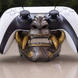 Könnte beinhalten: Ein weiß-schwarzer Videospiel-Controller liegt auf einer detaillierten Metallmaske mit goldenen Akzenten. Die Maske hat ein wildes Design mit offenem Mund, das einen auffälligen Kontrast zur schlanken Form des Controllers bildet. Thema: Gaming-Zubehör.