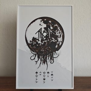 May include: A framed rectangular print featuring a large, dark, textured circular graphic resembling a moon with dripping elements at the bottom. Below the circle are several small, geometric symbols arranged in three vertical columns.