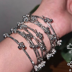May include: Silver bangles with floral and beaded detailing. The bangles are displayed on a wrist, highlighting their intricate design. The jewellery has a delicate, ornate appearance, suitable for special occasions or everyday wear.