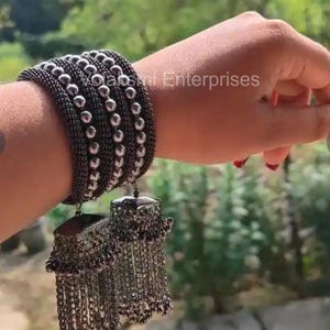 Può includere: Un set di braccialetti in metallo nero con perline color argento e orecchini pendenti. Gli orecchini hanno una parte superiore rettangolare con catenine e perline pendenti. I gioielli sono indossati su un polso.