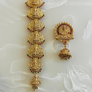 May include: Gold-coloured jewellery set with intricate designs. The set includes a long, vertical piece with multiple decorative elements and a matching earring. Each piece is adorned with red and green accents and a bell-shaped drop.
