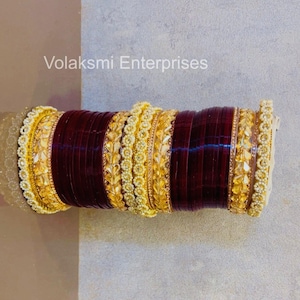 May include: A stack of bangles featuring deep maroon bands alternating with gold-toned textured bands and white beaded accents. The text Volaksmi Enterprises is visible above the stack. The set includes multiple thin maroon rings and decorative gold pieces.