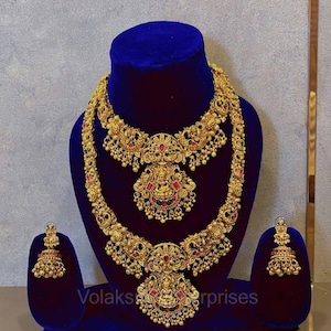 Puede incluir: Un conjunto de joyería en tono dorado que incluye dos collares superpuestos y un par de pendientes jhumka a juego. Las piezas presentan detalles intrincados con patrones en relieve, pequeñas cuentas colgantes y piedras rojas y verdes. El texto Volaks Enterprises es visible.