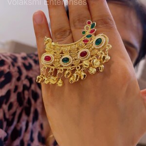 Op de afbeelding: Een goudkleurige statement ring met een sierlijk halvemaanvormig ontwerp met gedetailleerd metaalwerk, ingelegd met ovale rode en groene stenen, kleine witte kralen en een reeks bungelende goudkleurige belletjes langs de onderrand.