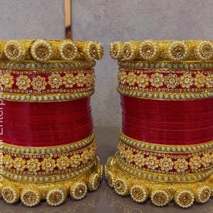 May include: Two sets of stacked bangles featuring a vibrant red base color. Each set is adorned with intricate gold-toned floral patterns, rows of small white beads, and a prominent border of larger, textured gold spheres along the top and bottom edges.