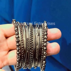 May include: A set of gunmetal-toned bangles featuring various textures, including smooth bands, beaded edges, and intricate etched patterns. The text Volatsmi Enterprises is visible across the top of the metallic bracelets.