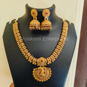 Puede incluir: Juego de collar y pendientes dorados exhibido en un maniquí negro. El collar presenta un colgante central con una piedra roja y detalles intrincados. Los pendientes a juego con piedras rojas y gotas en forma de campana completan el conjunto. El texto "Volaksmi Enterprises" es visible.