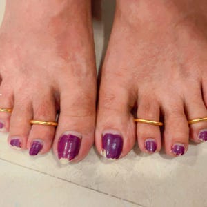 Pode incluir: Quatro anéis de dedo do pé em metal dourado liso, usados no segundo dedo de ambos os pés. As unhas dos pés estão pintadas com um esmalte roxo escuro. O texto Volaksmi Enterprises é visível verticalmente no lado esquerdo perto dos dedos.