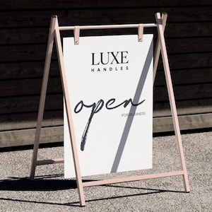 Może przedstawiać: Tablica w kształcie litery A z jasnoróżową ramą i białym znakiem. Znak głosi "LUXE HANDLES" na górze, ze słowem "open" w odręcznej czcionce i "FOR BUSINESS" poniżej. Znak znajduje się na szarej powierzchni.