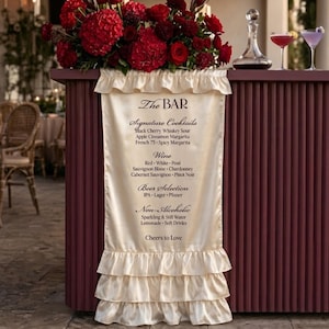 May include: A cream-coloured satin bar menu sign with ruffled edges, featuring black text listing The BAR, Signature Cocktails, Wine, Beer Selection, and Non-Alcoholic drinks, topped with a large arrangement of deep red roses and greenery.