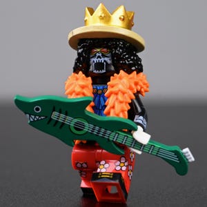 Puede incluir: Una figura de Lego de un personaje esqueleto con una corona dorada y piel naranja. La figura sostiene una guitarra verde con forma de tiburón. El personaje lleva pantalones rojos con diseños florales.