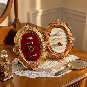 May include: Two ornate, gold-coloured ring boxes, open to display rings. One box has a red velvet interior, the other a cream-coloured interior. The boxes are oval-shaped with decorative frames. A small perfume bottle and a hand mirror are also visible.