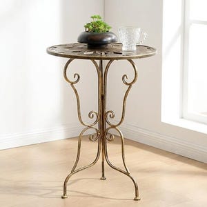 Può includere: Un tavolino decorativo in stile antico con un piano rotondo a motivi e gambe ornate. Il tavolo è in metallo con finitura bronzo. Una piccola pianta in vaso e un bicchiere trasparente sono sul tavolo. Il tavolo è alto circa 61 cm.