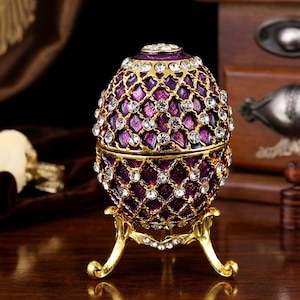 Puede incluir: Una caja de baratijas decorativa en forma de huevo púrpura con un diseño de filigrana dorada y brillantes detalles de cristal. El huevo se apoya en un soporte dorado, creando una exhibición elegante. El huevo mide aproximadamente 7.6 cm de alto.