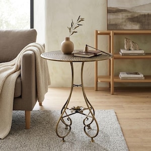 Peut inclure: Une table d'appoint ronde en métal avec un plateau texturé et une base incurvée ornée d'un motif central en forme de fleur. La table présente une finition couleur bronze et repose sur trois pieds en volutes. Un vase beige et un livre fermé.