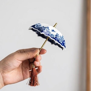 Op de afbeelding: Een kleine decoratieve paraplu met een wit en blauw bloemmotief. Het heeft een goudkleurige handgreep en een bruine kwast. Het patroon doet denken aan traditionele porseleinen ontwerpen.
