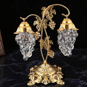 May include: A decorative gold-colored centerpiece featuring two clear glass grape-shaped containers. The containers are suspended from a gold vine design with leaves and grapes, resting on a detailed gold base. The overall design is ornate and elegant.