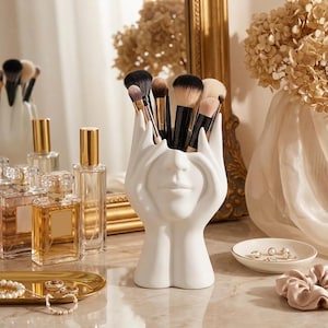 Peut inclure: Un porte-pinceaux de maquillage en céramique blanche en forme de visage avec des mains. Le support est rempli de divers pinceaux de maquillage. Des flacons de parfum, un plateau doré avec des bijoux et une petite coupelle avec des bijoux sont également visibles.