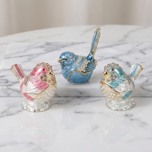 Puede incluir: Tres figuras decorativas de pájaros en rosa, azul y turquesa. Cada pájaro está adornado con pedrería y detalles dorados. Los pájaros están colocados sobre una superficie de mármol blanco, mostrando sus intrincados detalles y colores vibrantes.