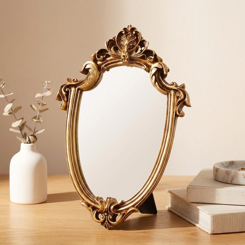 May include: A gold-colored, ornate vanity mirror with an oval shape. The mirror has a decorative, detailed frame with a floral design at the top and bottom. The mirror is standing on a wooden surface.