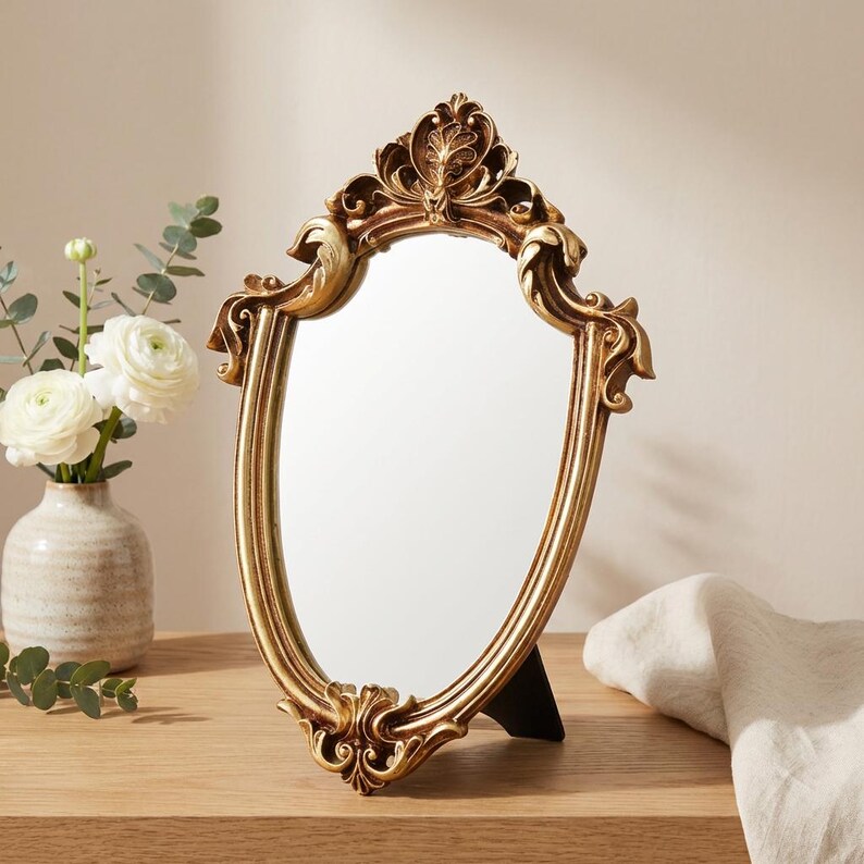 May include: A gold-colored, ornate vanity mirror with an oval shape. The mirror has a decorative, detailed frame with a floral design at the top and bottom. It stands on a black easel-style stand. The mirror is on a wooden surface.