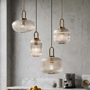 Può includere: Quattro lampade a sospensione in vetro trasparente con dettagli a coste e accenti metallici color oro sono appese al soffitto. Le luci hanno forme e dimensioni diverse, illuminando un ambiente cucina. Un piano di lavoro in legno e una ciotola di limoni sono in primo piano.