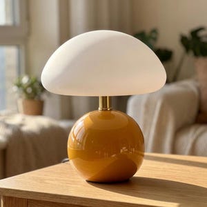 May include: A table lamp featuring a smooth, white, mushroom-shaped glass shade resting on a glossy, spherical mustard yellow base. The two sections are connected by a short, cylindrical metallic gold-toned neck, creating a balanced, rounded silhouette.