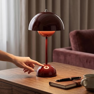 Può includere: Una lampada da tavolo bordeaux con un paralume a forma di cupola e un gambo sottile. La lampada si accende con una base sensibile al tocco. La lampada è su un tavolo di legno con un taccuino, una penna e una tazza.