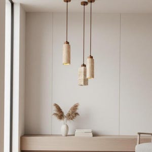 May include: A set of three hanging pendant lights featuring cylindrical and rectangular shades made of beige travertine stone. Each light is suspended by a thin dark brown cord from the ceiling, emitting a warm glow from the bottom opening.
