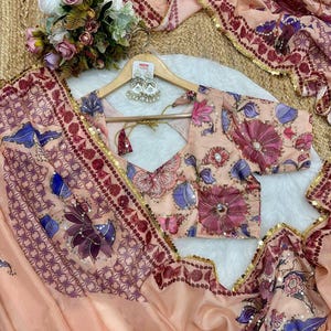 May include: Peach-colored floral saree set with a matching blouse. The saree features intricate floral patterns in shades of purple, blue, and pink. The blouse has a V-neck and short sleeves. The set is embellished with gold sequins.