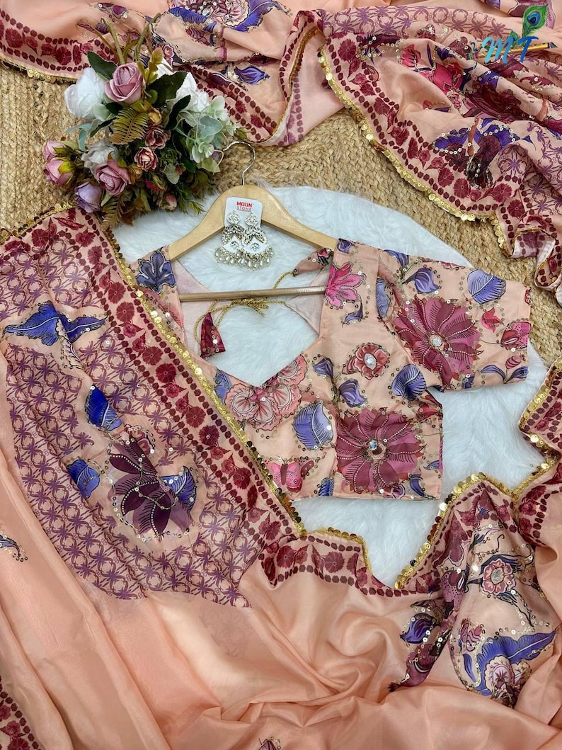 May include: Peach-colored floral saree set with a matching blouse. The saree features intricate floral patterns in shades of purple, blue, and pink. The blouse has a V-neck and short sleeves. The set is embellished with gold sequins.