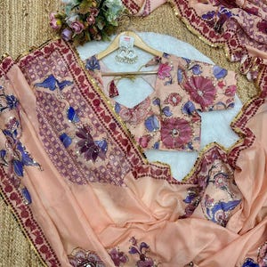 May include: A peach-colored saree and blouse set with floral and geometric patterns in shades of purple, blue, and pink. The saree has a decorative border with gold sequins. The blouse is on a wooden hanger with a pair of earrings.