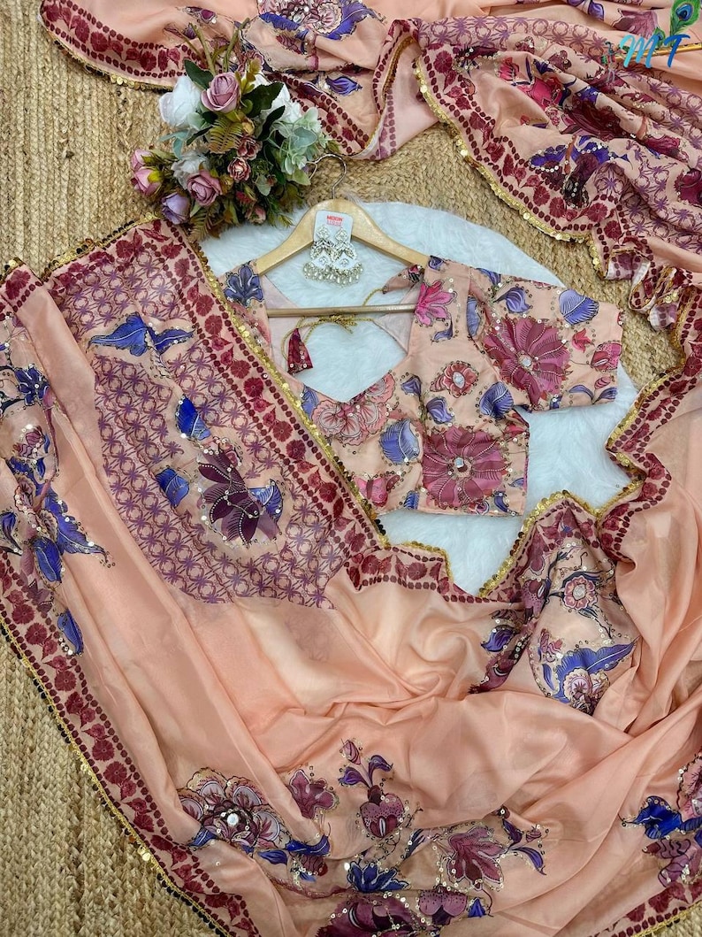 May include: A peach-colored saree and blouse set with floral and geometric patterns in shades of purple, blue, and pink. The saree has a decorative border with gold sequins. The blouse is on a wooden hanger with a pair of earrings.