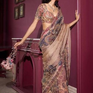 May include: A woman wearing a detailed, patterned saree with a matching blouse. The saree features intricate embroidery in shades of pink, gold, and blue. She is also wearing a necklace. The background includes a decorative cabinet and framed artwork.