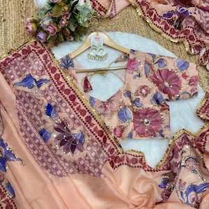 May include: A peach-colored saree and blouse set with floral and geometric patterns. The saree features a border with gold sequins and a matching blouse. The set includes a pair of silver earrings on a wooden hanger. A bouquet of flowers is also visible.
