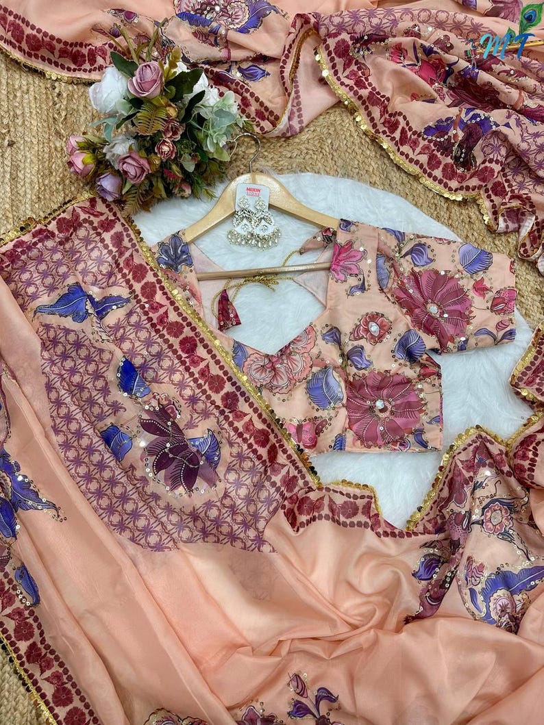 May include: A peach-colored saree and blouse set with floral and geometric patterns. The saree features a border with gold sequins and a matching blouse. The set includes a pair of silver earrings on a wooden hanger. A bouquet of flowers is also visible.
