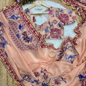 May include: A peach-colored saree and blouse set with floral embroidery in shades of purple, blue, and pink. The saree has a decorative border with gold sequins. The blouse features a V-neck and short sleeves, with matching floral patterns.