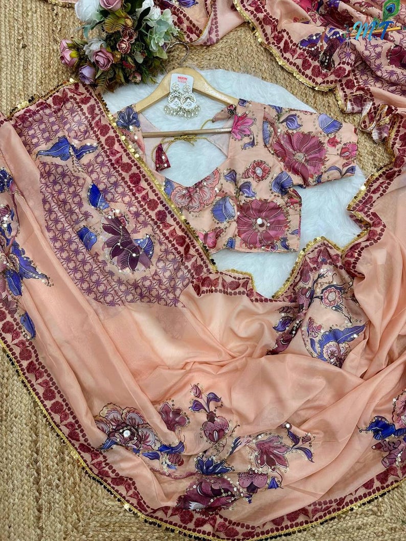May include: A peach-colored saree and blouse set with floral embroidery in shades of purple, blue, and pink. The saree has a decorative border with gold sequins. The blouse features a V-neck and short sleeves, with matching floral patterns.