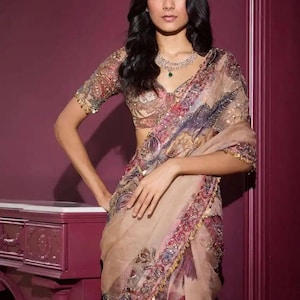 May include: A beige saree with intricate floral and paisley patterns in shades of red, purple, and blue. The saree features a decorative border with gold embellishments. The model is wearing a matching blouse and a diamond necklace with an emerald pendant.