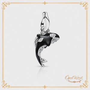May include: A silver charm featuring a black and white orca whale design. The whale has a detailed, curved shape with sparkling accents. The charm is suspended from a silver loop, suitable for a bracelet or necklace. The charm is a jewelry item.
