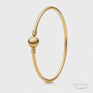 Pode incluir: Uma pulseira de bracelete dourada com um berloque esférico. O berloque é gravado com texto e está posicionado no centro da pulseira. A pulseira tem um acabamento liso e polido e um design simples e elegante.